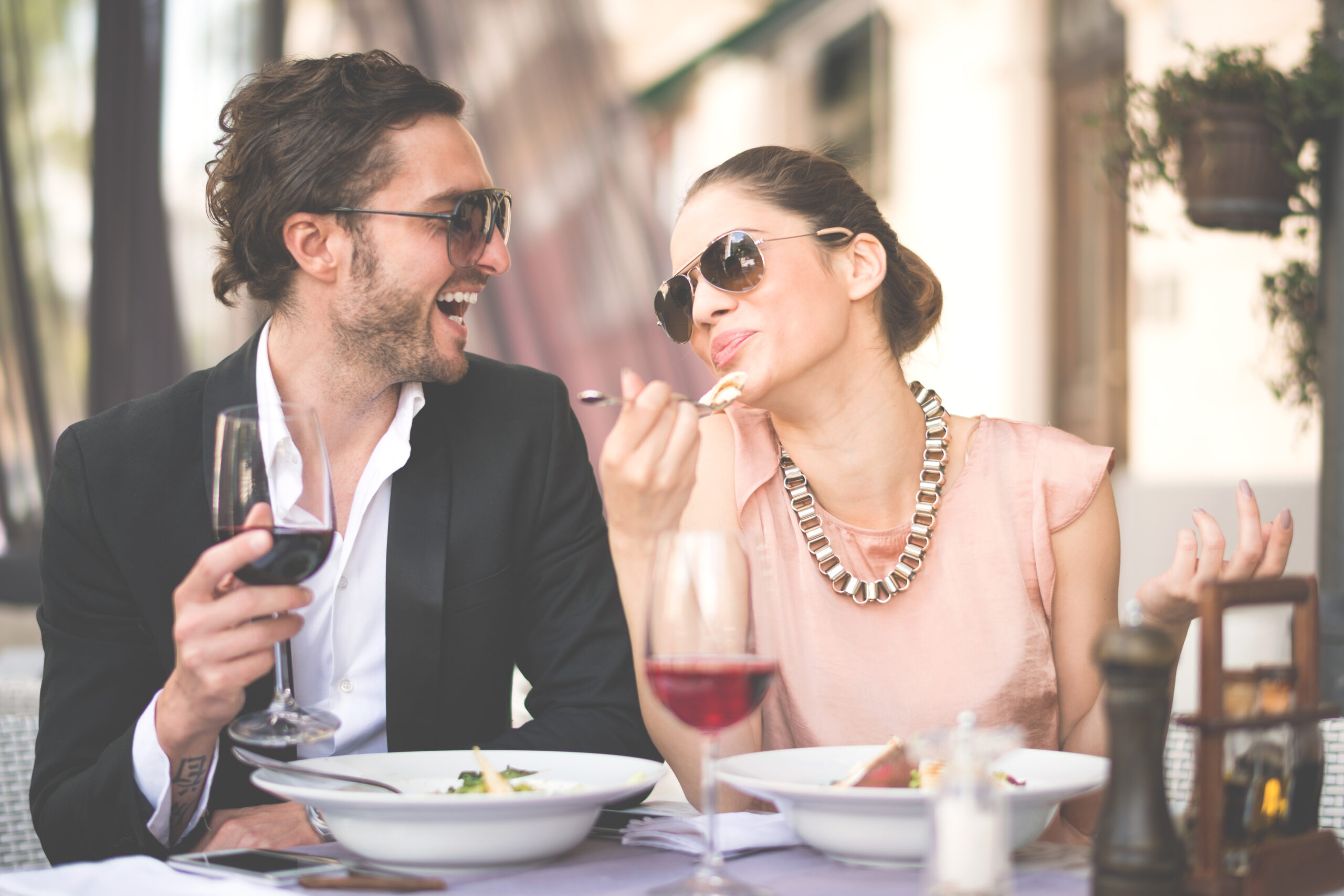 Nicely dressed couple with sunglasses sitting in restaurant and having a meal. Man is holding his wine glass and woman is about to take a bite. They are looking at each other and smiling.