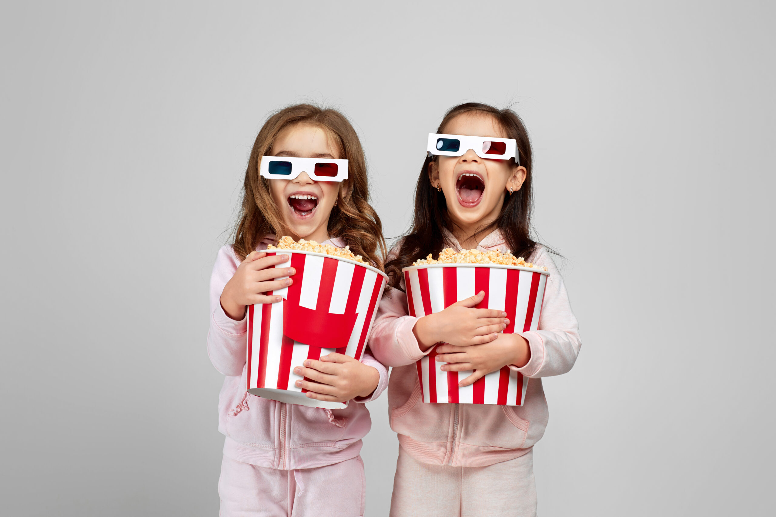 two beautiful surprised caucasian little girls in red-blue 3d glasses holding popcorn buckets and watching movie