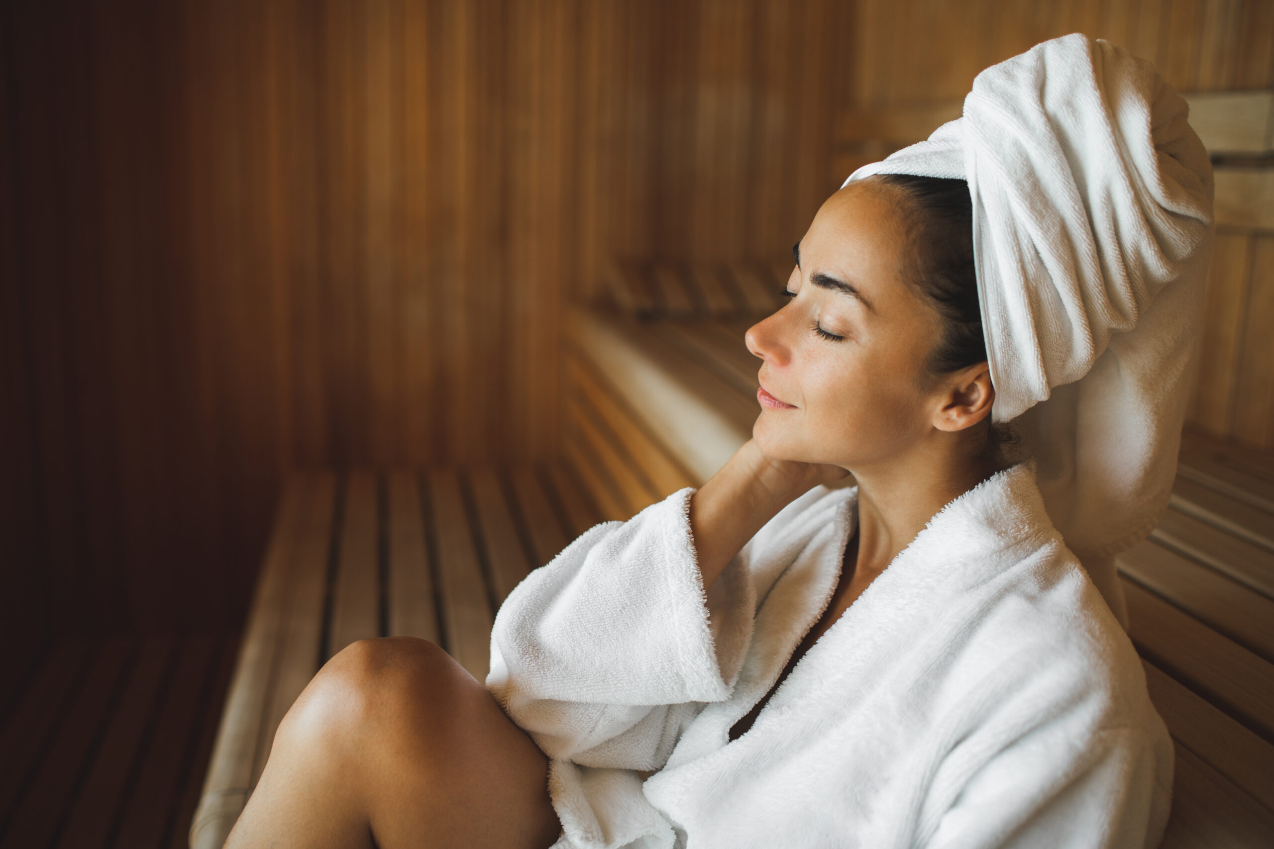 Young beautiful slim woman in wooden dry sauna in spa centr in white bathrobe with towel on her head. Relaxation wellness and beauty concept. Finnish sauna for strengthening the body's immune system, removing toxins, resting body and mind.