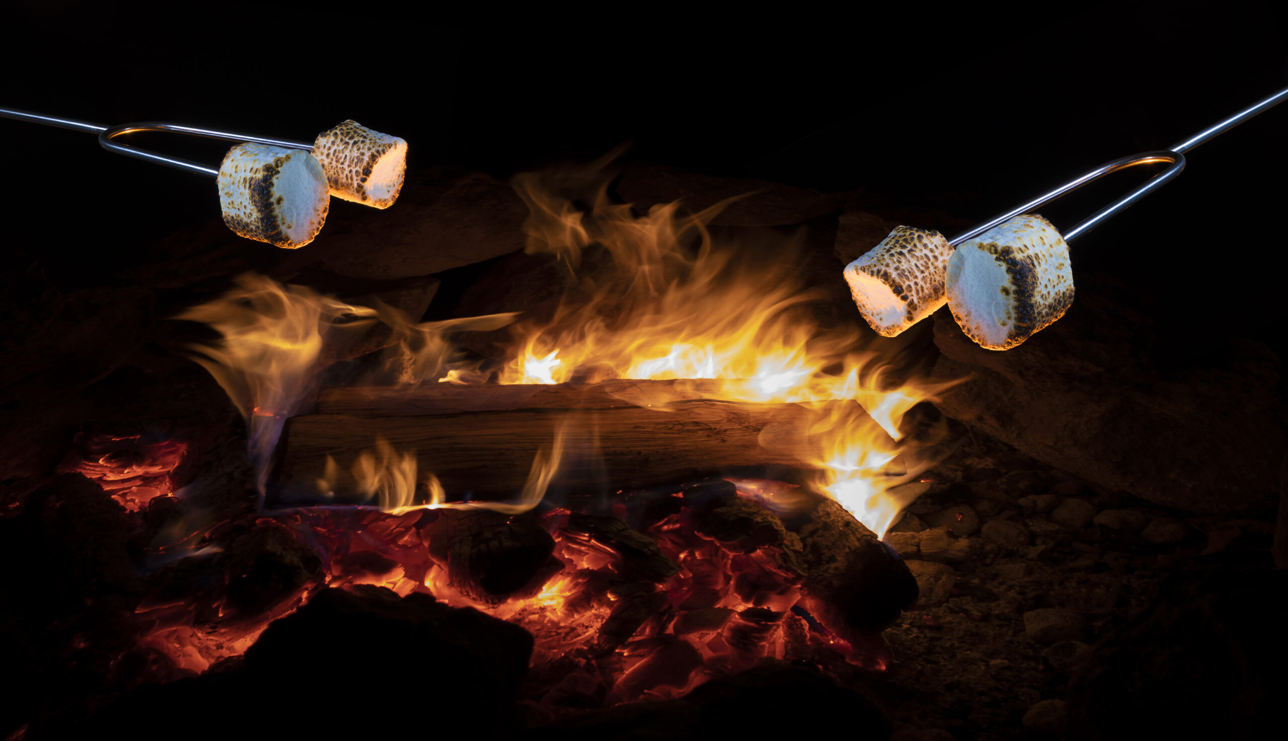 Pair of skewers with marshmallows being roasted over a warm open fire outdoors at night